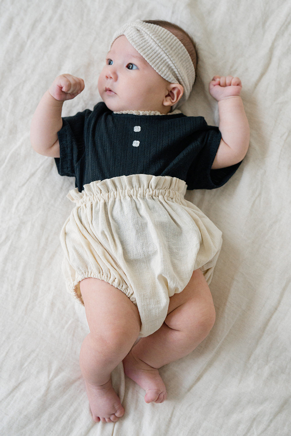 Baby lying on a blanket wearing Avauma Berry Puff Bloomer in Soft Ivory, styled with a black top and ribbed beige headband