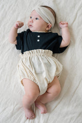 Baby lying on a blanket wearing Avauma Berry Puff Bloomer in Soft Ivory, styled with a black top and ribbed beige headband