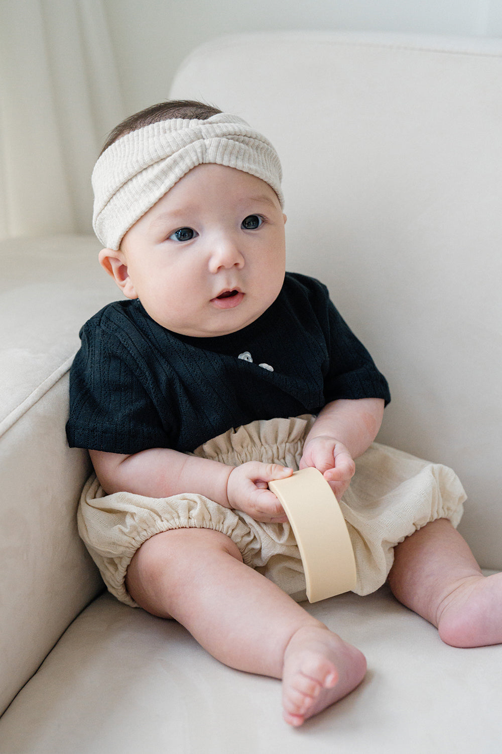 Baby sitting on a sofa wearing Avauma Berry Puff Bloomer in Soft Ivory with ribbed headband and black ribbed top