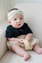 Baby sitting on a sofa wearing Avauma Berry Puff Bloomer in Soft Ivory with ribbed headband and black ribbed top