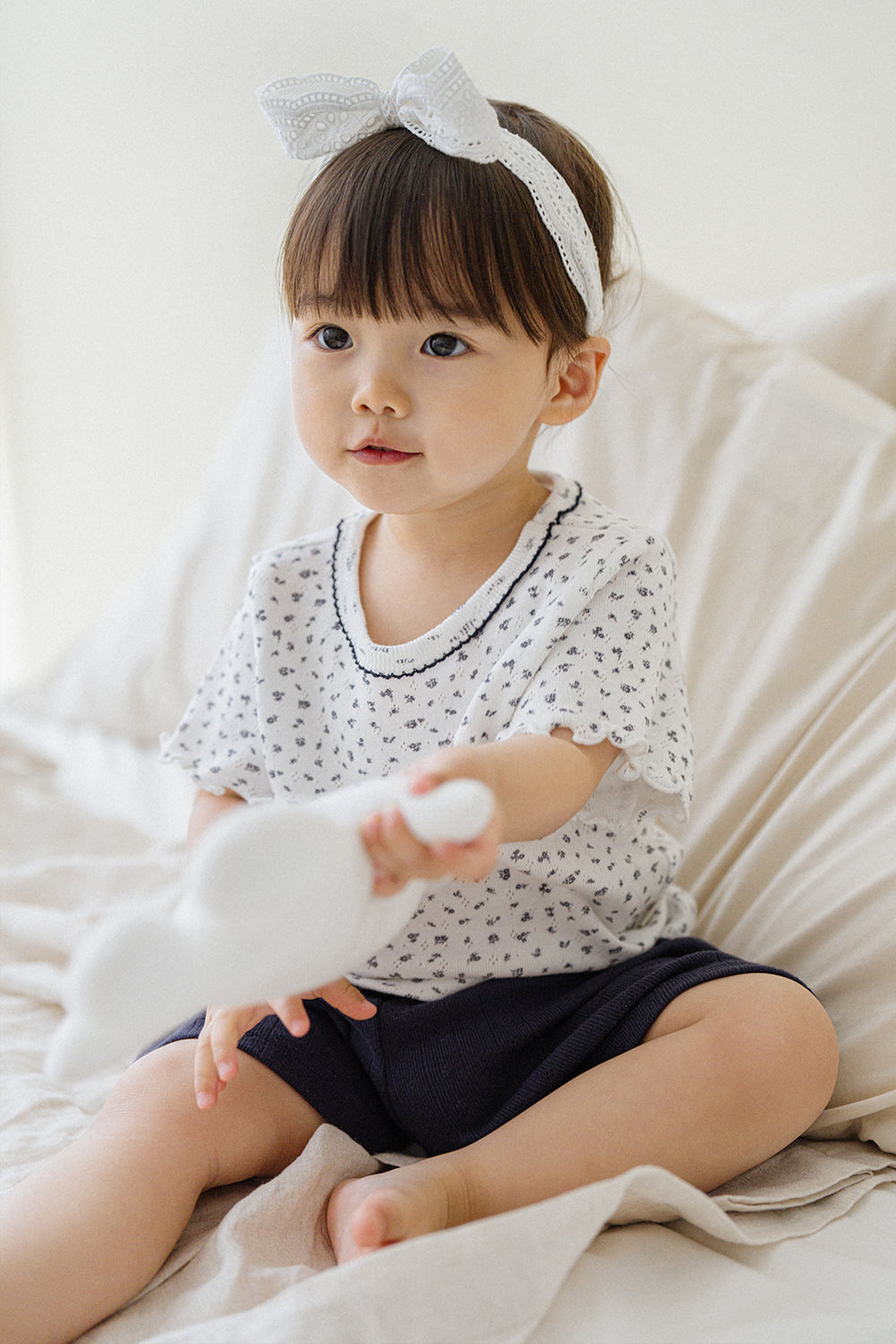 Avauma breeze garden baby setup indigo whisper - girl with headband holding plush toy in white floral top and navy shorts on bed

