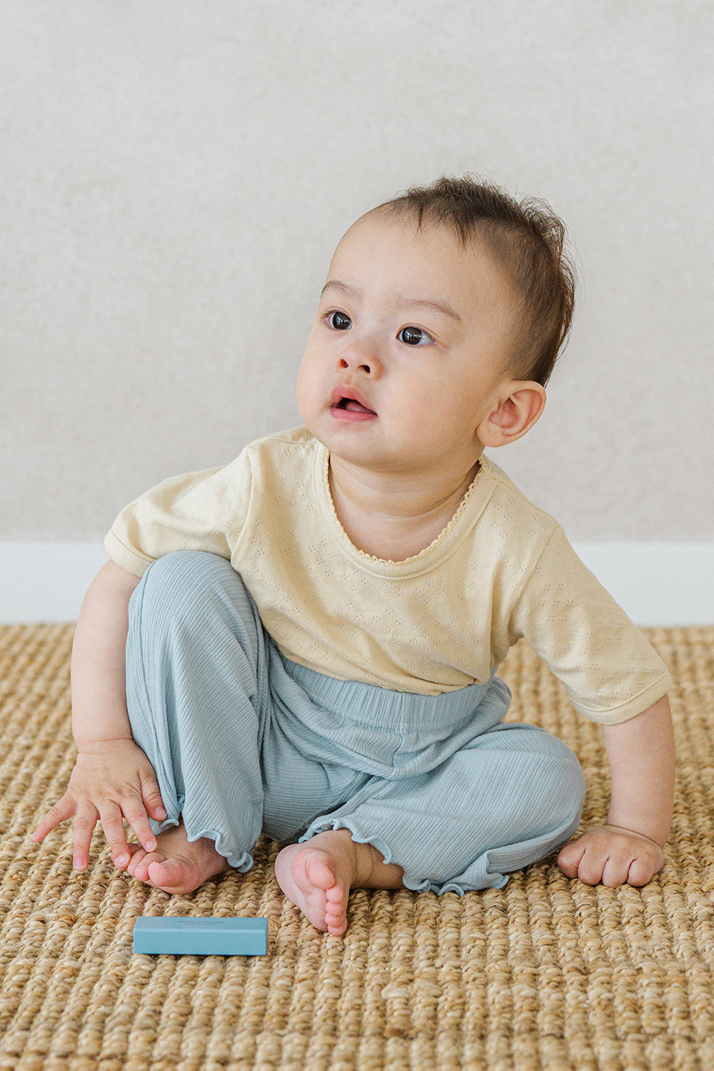 Infant in Avauma oat beige lace-trimmed short sleeve tee and blue ribbed pants, playing on carpet

