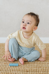 Infant in Avauma oat beige lace-trimmed short sleeve tee and blue ribbed pants, playing on carpet

