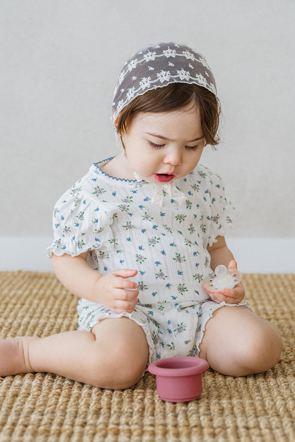 Cute baby in Avauma Petit Ruffle Bluebell summer set, playing with a pacifier on the carpet.


