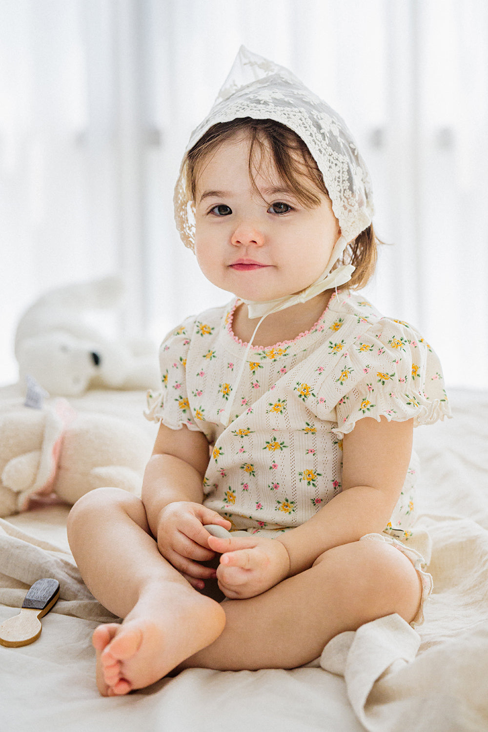 Smiling baby girl wearing lace bonnet and Avauma Petite Ruffle Short Sleeve Set - Honey Daisy, sitting on a soft blanket.

