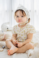 Smiling baby girl wearing lace bonnet and Avauma Petite Ruffle Short Sleeve Set - Honey Daisy, sitting on a soft blanket.

