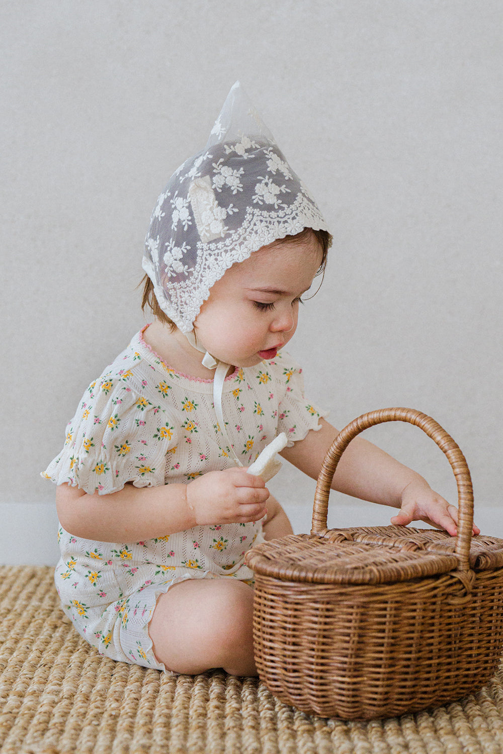 Baby wearing Avauma Petite Ruffle Short Sleeve Set - Honey Daisy and lace bonnet, sitting on a rug and reaching for a wicker basket.

