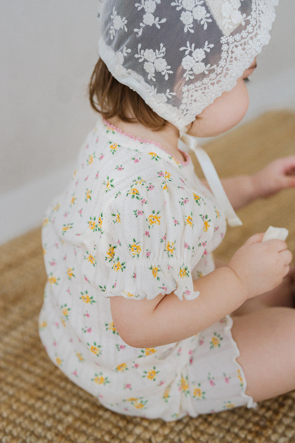Baby wearing a lace bonnet and Avauma Petite Ruffle Short Sleeve Set in Honey Daisy, side view close-up showing yellow daisy floral pattern.

