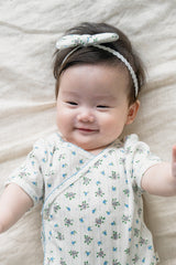 Smiling baby girl wearing Avauma Sky Garden Petite Garden Headband with blue floral print, lying on a beige blanket