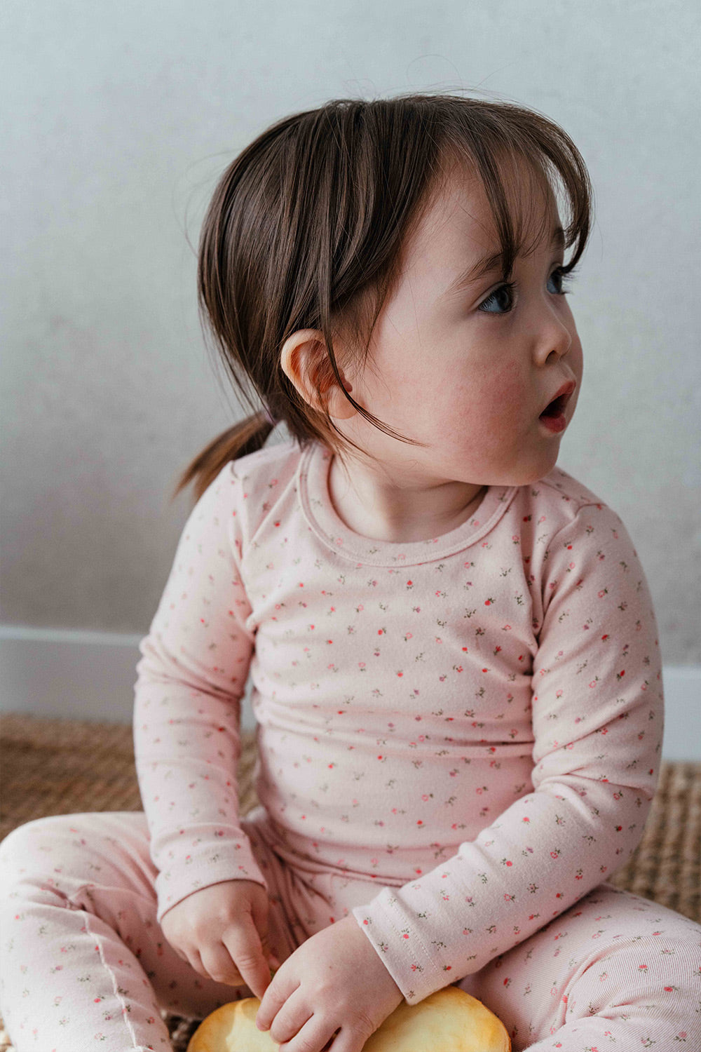 Toddler girl wearing Avauma Floral Melody Long Sleeve Baby Pajamas - Powder Pink, sitting and looking to the side in a cozy floral print pajama set