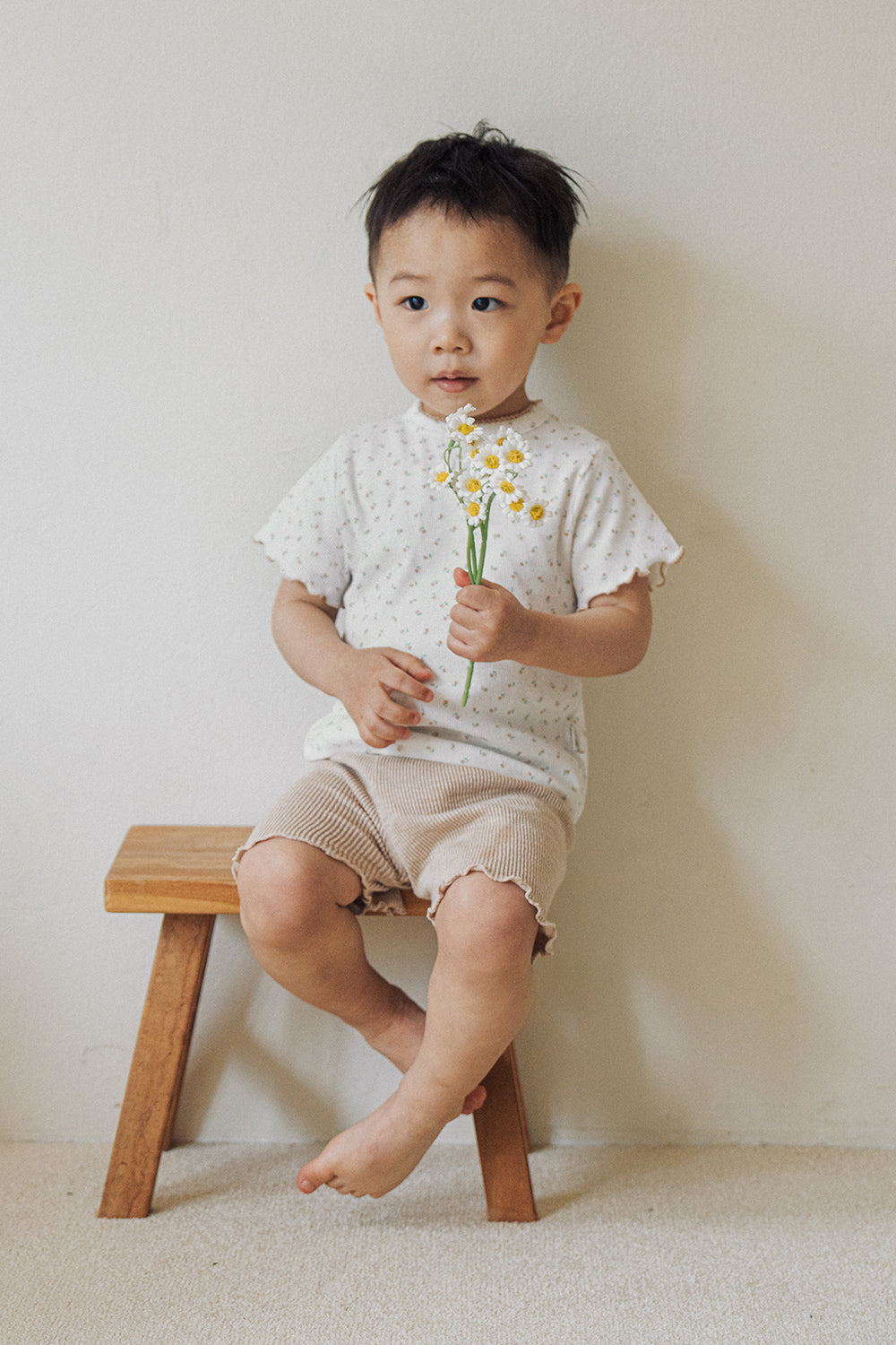 A toddler boy sitting on a wooden stool wearing Avauma honey beige floral t-shirt and shorts.