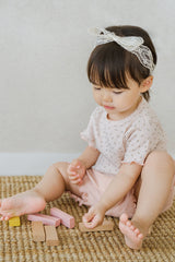 Toddler girl sitting and playing with wooden blocks, wearing rosy blush baby summer set with pink shorts and floral top.

