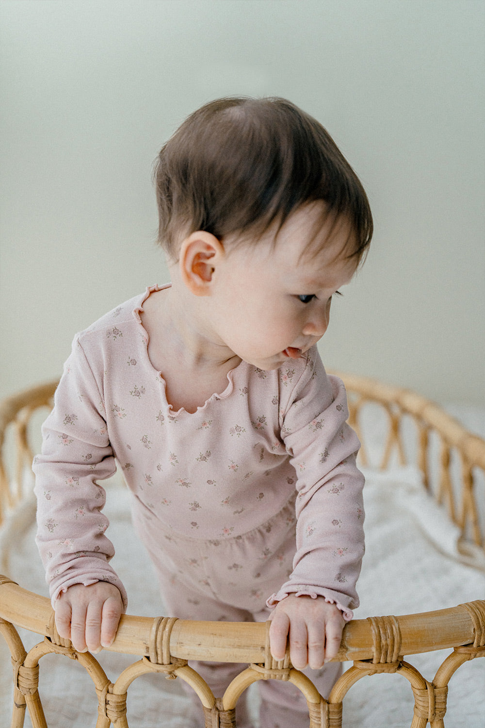 Baby standing in a crib, dressed in Avauma Fluid Blossom Long Sleeve Baby Pajamas - Strawberry Milk, highlighting the cozy pink pajama set with delicate floral designs.